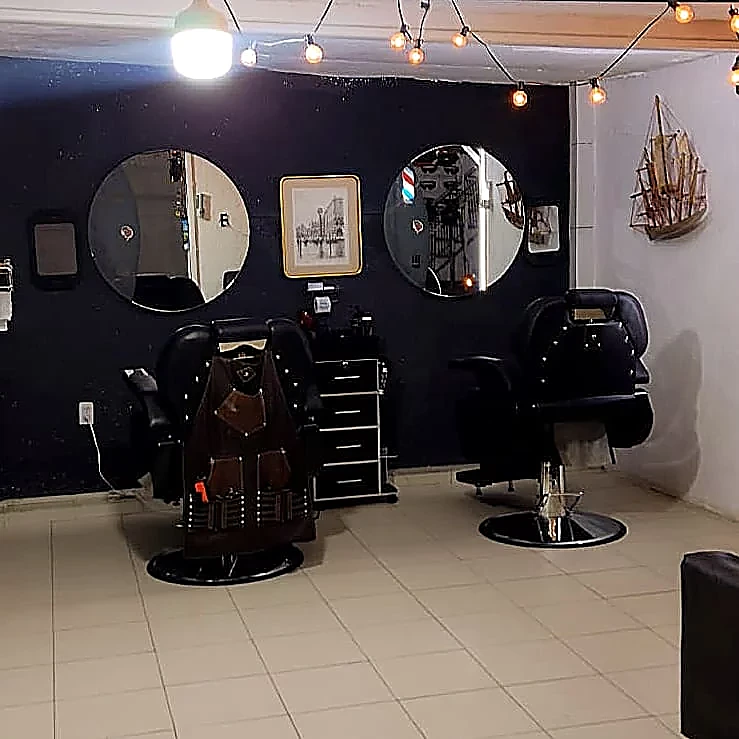 Dos espejos redondos sobre pared negra, sillones de barbero, una serie de foquitos colgados y un barco de madera en la repisa