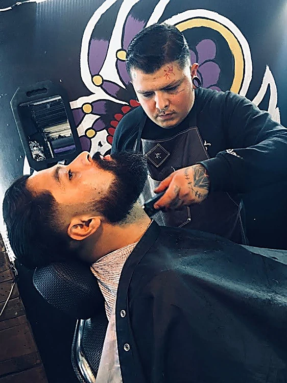 Barbero perfilando con máquina la barba de un cliente recostado en el sillón, con el mural morado al fondo