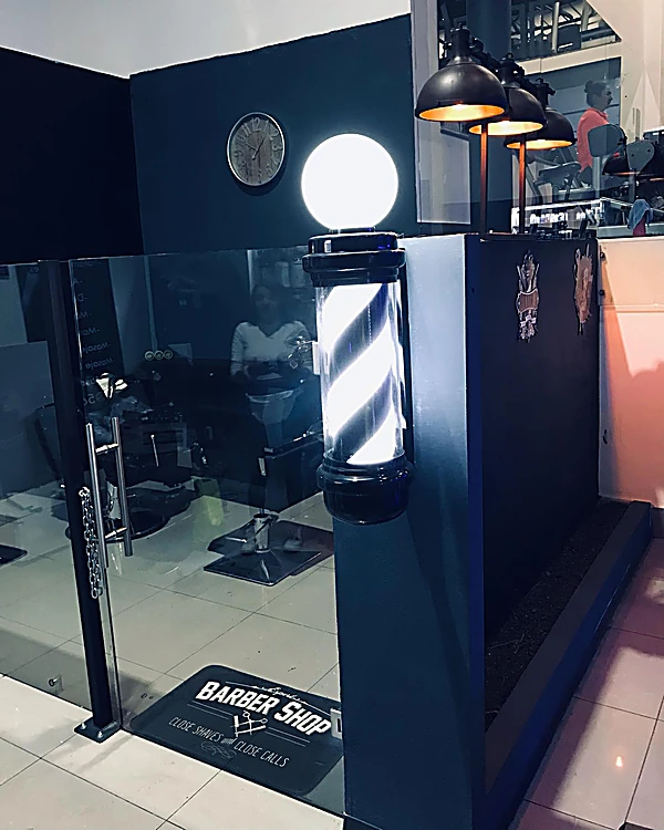 Poste de barbero rojo, blanco y azul con globo de luz junto a la entrada, tapete que dice Barber Shop y lámparas negras